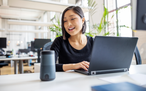 Business professional working on laptop and talking to a virtual assistant via smart speaker