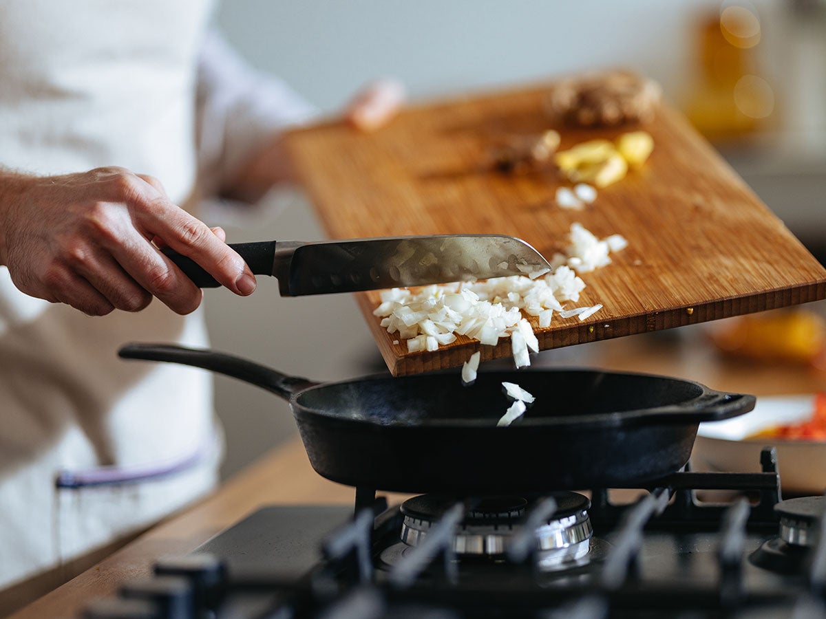 A chef scraping diced onions off a cutting board into a skillet on a gas stove