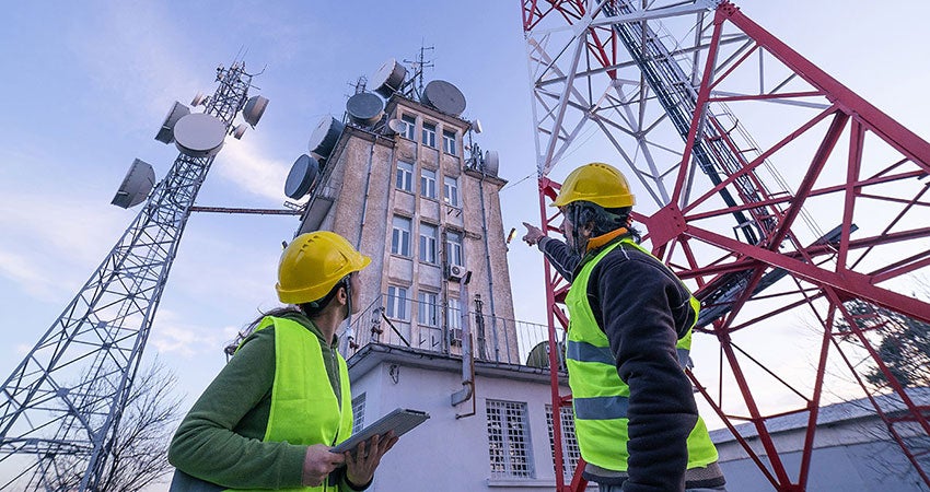 Two workers in safety gear inspecting telecom towers near a building with antennas