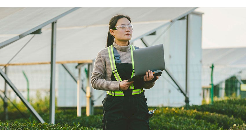 Person in safety gear holding a laptop in a greenhouse