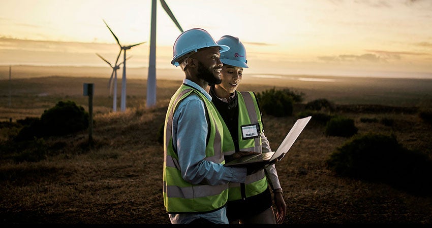 Two workers in safety vests and helmets using a laptop near wind turbines at sunset