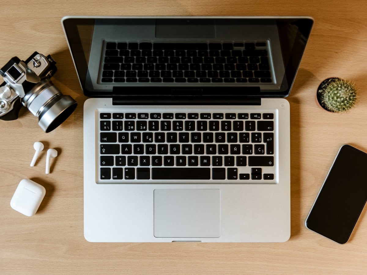 Overhead view of a laptop on a desk sitting next to a cellphone, wireless headphones and a camera