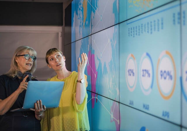 Professionals review data on a laptop and on a larger wall display