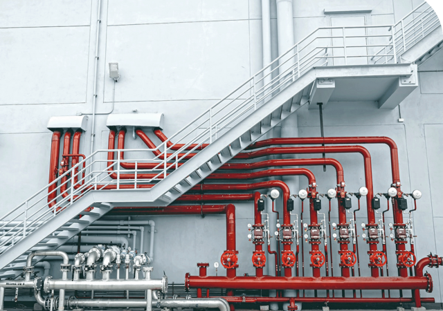 Red industrial pipes and valves mounted on a gray wall beneath a metal staircase.