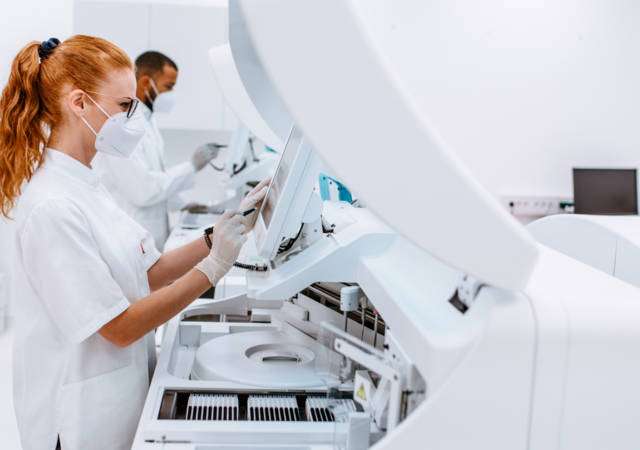 Two laboratory professionals wearing white lab coats, face masks, and gloves work in a modern, sterile lab. The woman in the foreground interacts with a large automated diagnostic machine via a touchscreen, while the man in the background operates another piece of equipment. The setting highlights advanced medical technology and strict hygiene protocols.