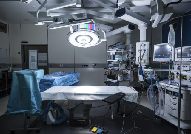 A modern operating room featuring a central surgical table covered with a white sterile sheet, surrounded by advanced medical equipment including patient monitors, anesthesia machines, and illuminated overhead surgical lights. Blue sterile drapes cover nearby instrument tables, and IV stands, tubing, and foot pedals are positioned for immediate use. The room is clean, organized, and designed for precision and safety in surgical procedures.
