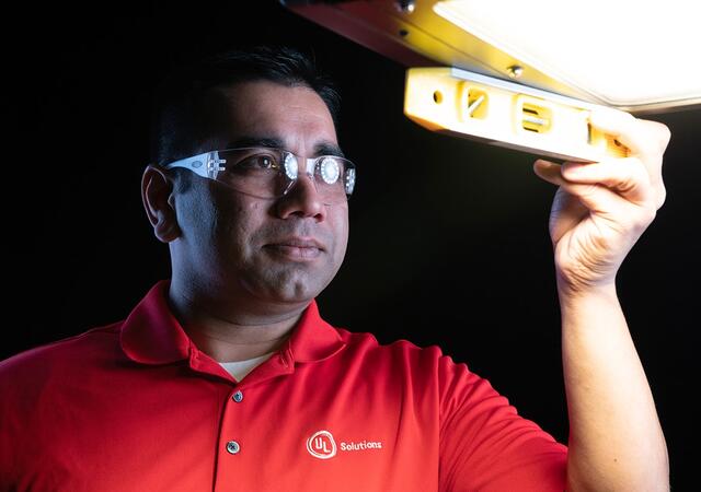 Person in a red UL Solutions shirt holding a yellow level under a bright overhead light.