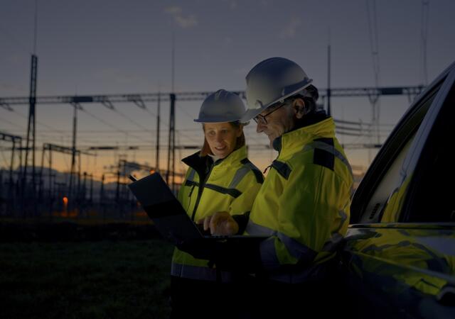 Two engineers looking at reports while standing next to a power grid at dawn