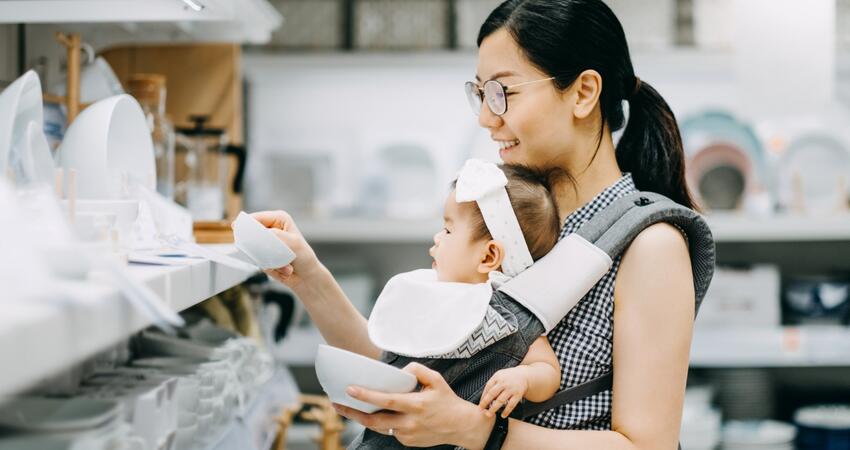 A woman wearing an infant in a soft carrier shops for dishes in a retail store.