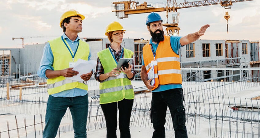Three construction workers in safety vests and helmets reviewing plans on a building site
