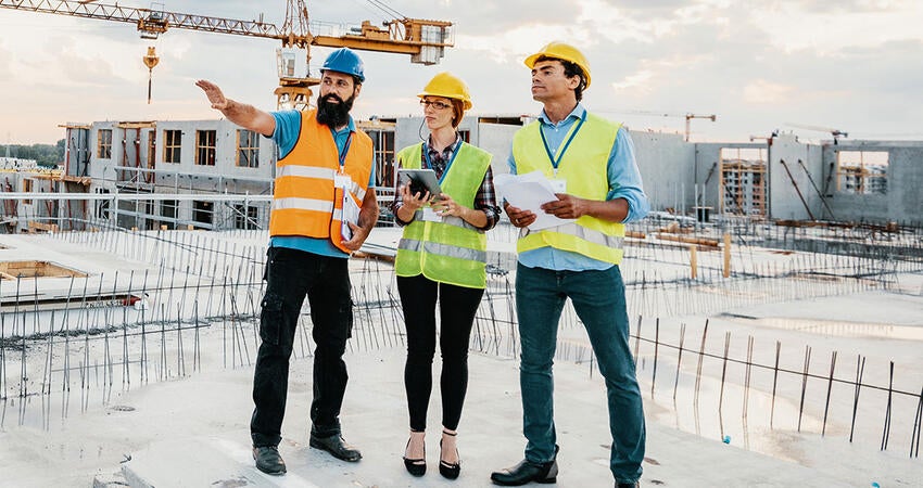 Three construction workers in safety vests and helmets reviewing plans on a building site.