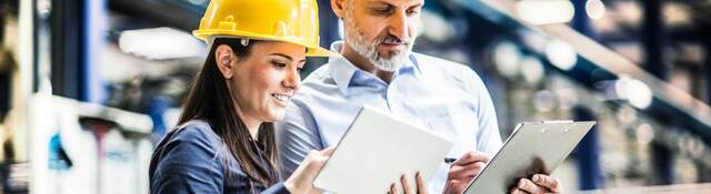 Two workers in yellow hardhats comparing notes on their tablet and clipboard 