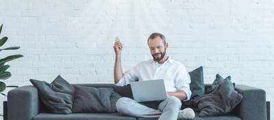 Bearded white man in light jeans and white sneakers on modern gray sofa with air conditioner on wall