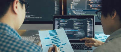 Two engineers examining a process flow on paper with coding on computer screens in the background.