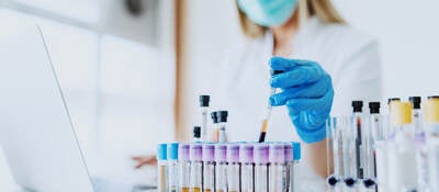 Woman wearing face mask in bright white lab observing vials while logging information on laptop