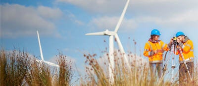 Two workers surveying in a field with wind turbines behind them