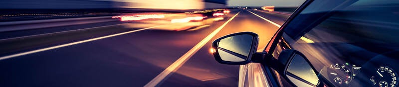 Silver car with side-view mirror driving on highway beside heavy duty truck at night