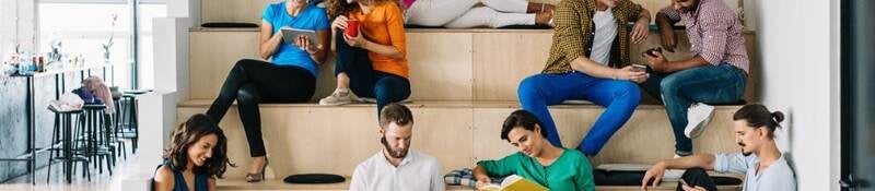 Group of young adults sitting on indoor tiered seating using their mobile phones and tablets.