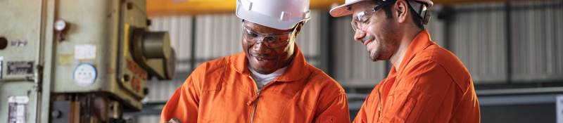 Two engineers in orange coveralls and white hardhats looking at a laptop