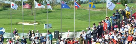 ULIC crowd watches golf event with flags flying in the background., UL International Crown Golf fans mill about the UL Fan expo during the four day event., Woman in red sweater looks through virtual reality glasses, UL Fan Expo visitors greeted by smiling volunteers and a big welcome sign in English and Korean. The volunteers, a man in a grey jacket and a woman in a red jacket smile and pose by a UL International Crown booth.