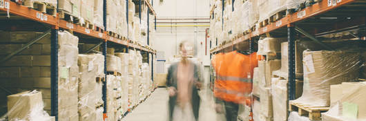 Two people walking through a warehouse of packages on shelves