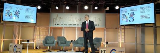 Tom Chapin, Ph.D, standing onstage with the 2019 Pritzker Forum on Global Cities banner in the center and two monitors showing the average number of batteries on any one person at any given time, to the left and right. Tom is wearing a blue suit with a red tie.