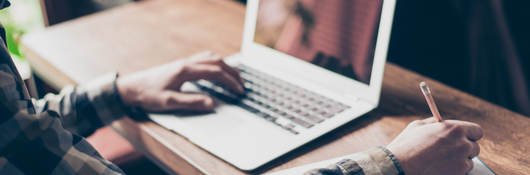 Man sitting at desk on laptop taking notes with pencil and paper