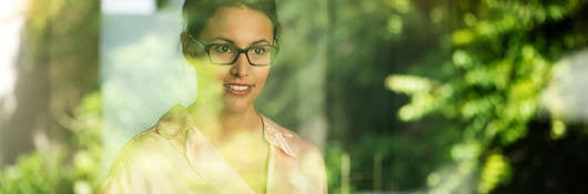 A woman stands at a window with reflected greenery.