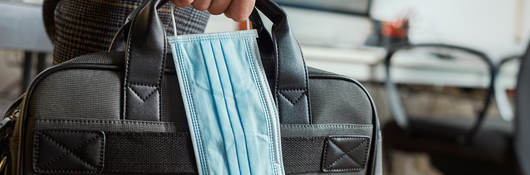 Man in an office holding a briefcase and surgical mask in his hand