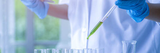 Laboratory worker with blue gloves holds a pipette with green liquid over a several test tubes