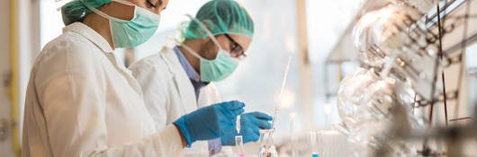 Two scientists wearing facemasks and examining chemical substances in test tubes in a laboratory.