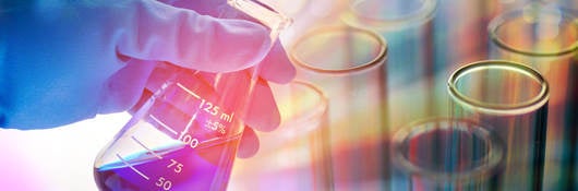 Rubber-gloved hand holding beaker containing blue liquid on a colorful background with test tubes