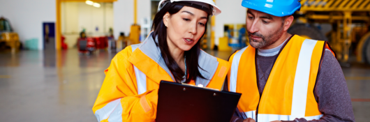 Two workers talking together over a clipboard while standing inside a large warehouse