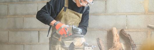 A carpenter, using proper safety equipment, rounds a piece of wood with an angle grinder.