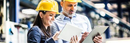 Two workers in yellow hardhats comparing notes on their tablet and clipboard 