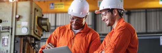 Two engineers in orange coveralls and white hardhats looking at a laptop
