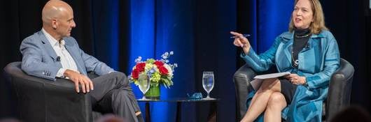 Jenny Scanlon talking while seated on main stage in blue dress. 