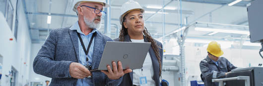 Two managers touring a factory floor while reviewing a laptop