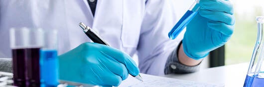 A chemist is analyzing sample and recording it in a notebook while in a laboratory with equipment and science experiments and glassware containing chemical liquids