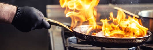 A chef wearing a protective glove while holding a frying pan that has flames coming off of it