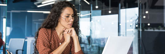 Person in a glass office, looking thoughtfully at a laptop