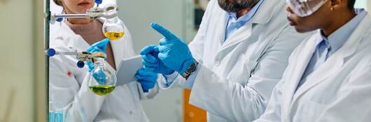 Three scientists wearing lab coats and gloves are engaged in a lively discussion about their research in a well-equipped laboratory, utilizing a laptop and a microscope for analysis