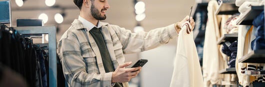  Giovane adulto che controlla il prezzo su smartphone in un negozio di abbigliamento - Foto stock Young man in a modern clothing store holds a cream sweater on a hanger, smiling as he checks his smartphone to compare prices, scan a qr code, or shop online
