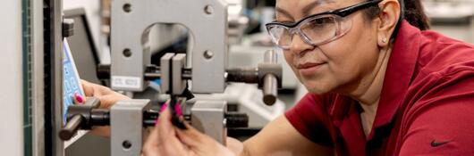 A laboratory professional wearing safety glasses adjusts a materials-testing machine.