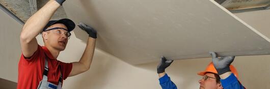 Two mean in hardhats install gypsum board to a ceiling