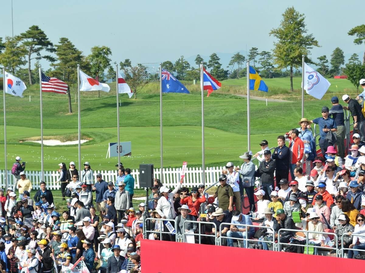 ULIC crowd watches golf event with flags flying in the background., UL International Crown Golf fans mill about the UL Fan expo during the four day event., Woman in red sweater looks through virtual reality glasses, UL Fan Expo visitors greeted by smiling volunteers and a big welcome sign in English and Korean. The volunteers, a man in a grey jacket and a woman in a red jacket smile and pose by a UL International Crown booth.