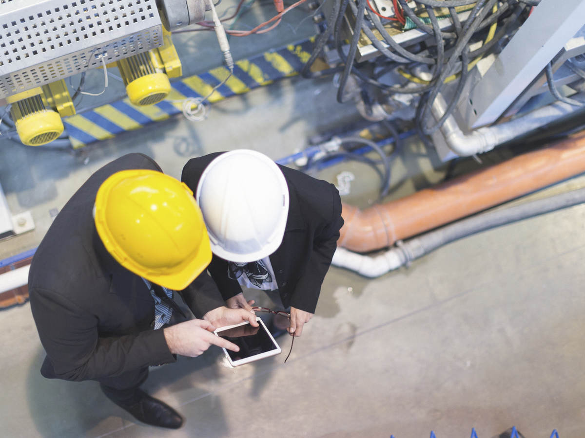 Two men in hard hats on a factory floor consulting over a tablet.