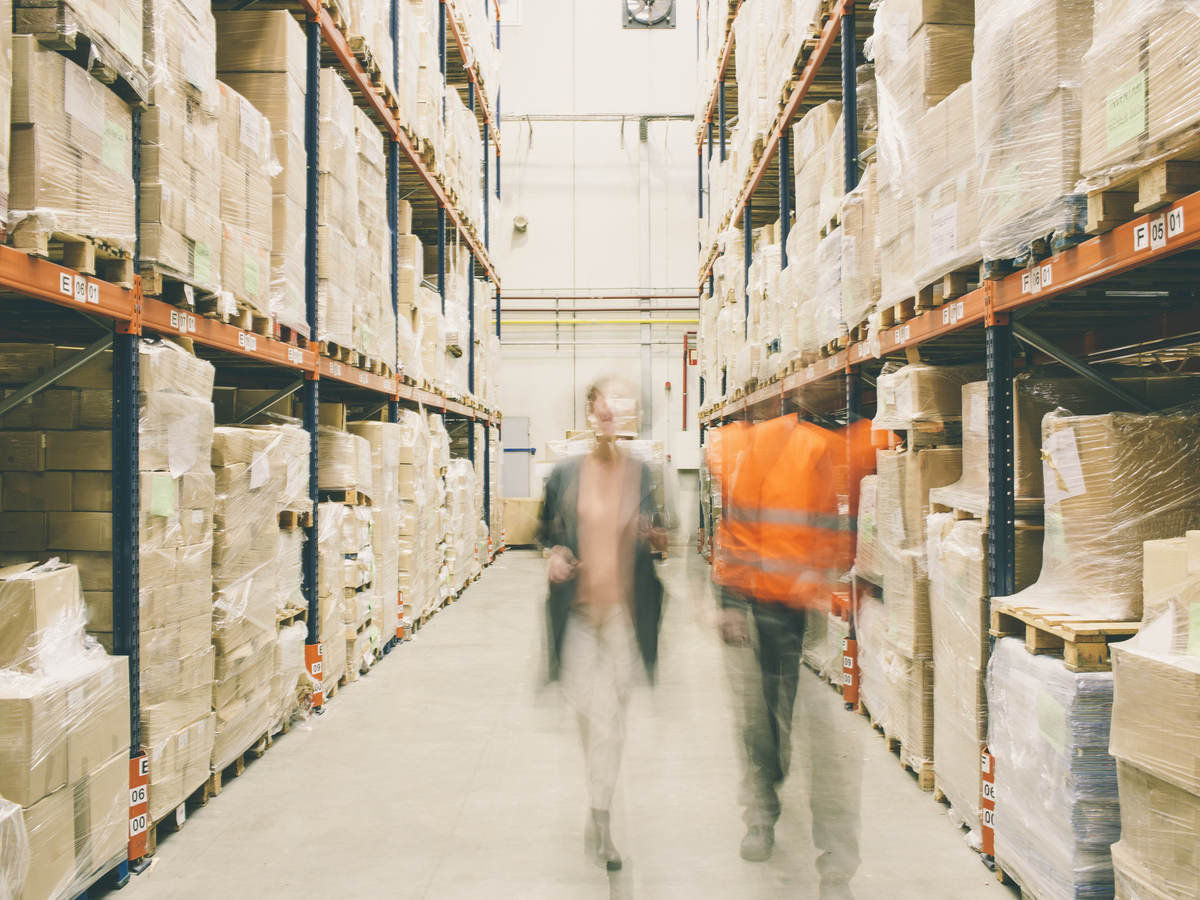 Two people walking through a warehouse of packages on shelves