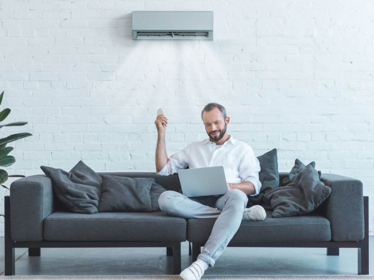 Bearded white man in light jeans and white sneakers on modern gray sofa with air conditioner on wall