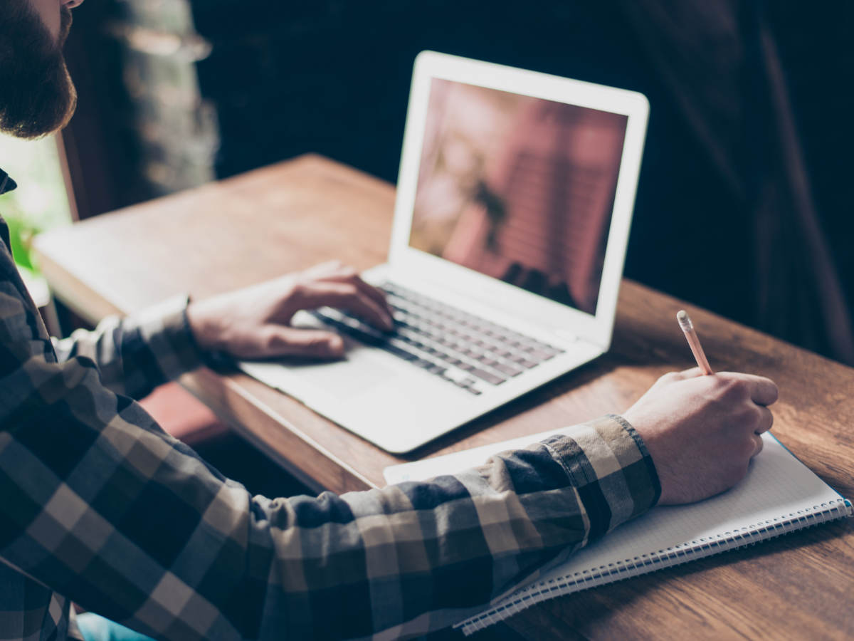 Man sitting at desk on laptop taking notes with pencil and paper